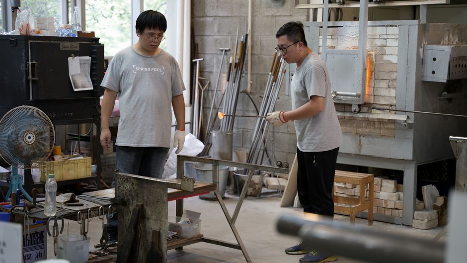 Two artisans intently engaged in glassblowing inside a lively workshop environment.