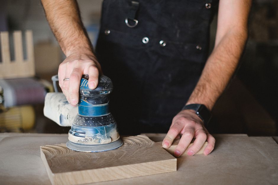Unrecognizable male carpenter using random orbital sander while working with wooden detail at workbench in professional workshop  on blurred background